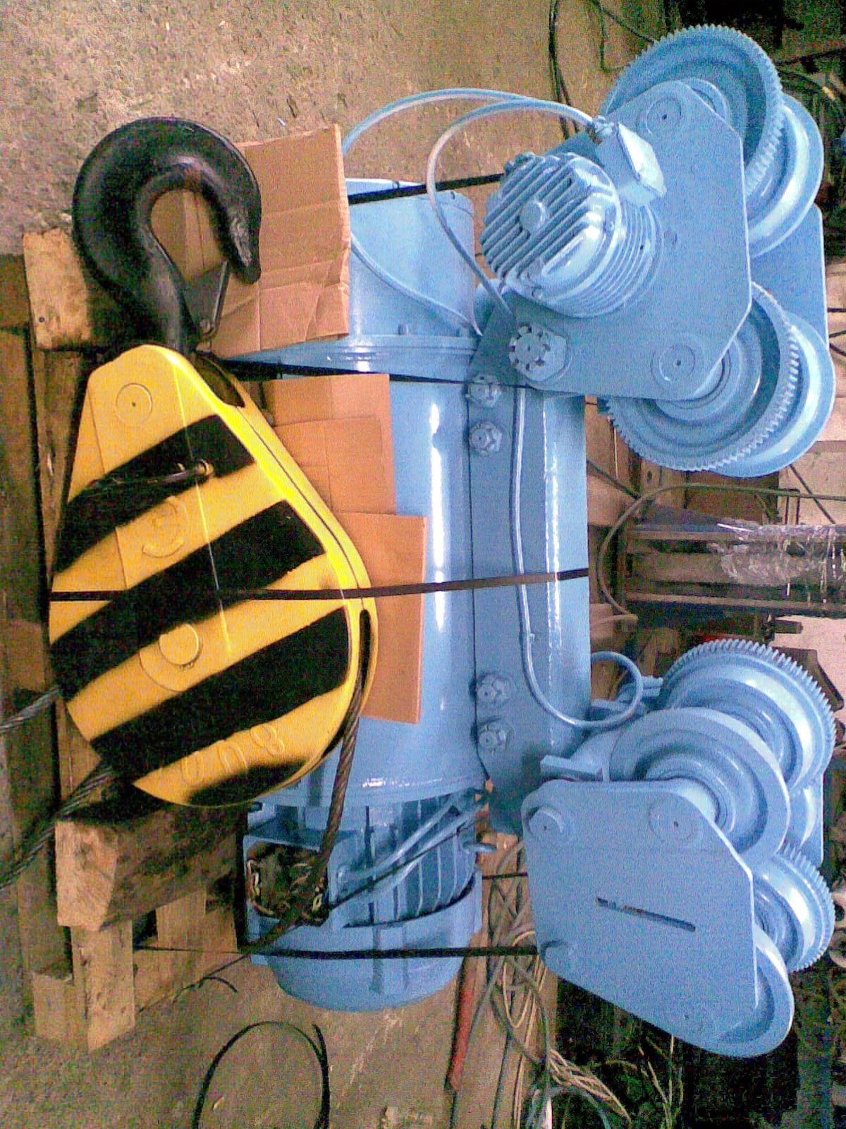 Elektrowciągnik 8t z żółto-czarnym hakiem na palecie, zabezpieczony kartonem, w odcieniach błękitu